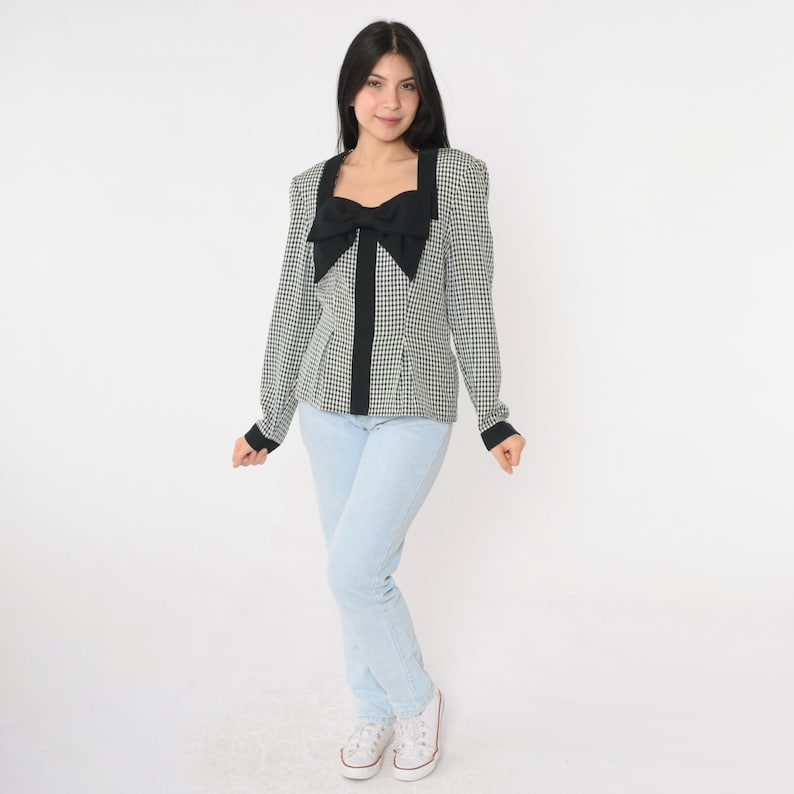 May include: A white and black gingham blouse with a large black bow at the neckline. The blouse has long sleeves and a fitted silhouette.