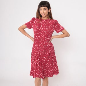 May include: A red dress with white polka dots. The dress has short sleeves, pleats down the front, and a pleated skirt. The model is wearing the dress and has their hands on their hips.