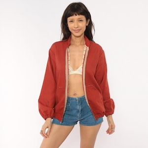 May include: A rust-colored zip-up jacket featuring long sleeves with elasticized cuffs, a high collar, and a front zipper closure. The interior lining displays a subtle grid pattern in neutral tones, visible along the open front edges of the garment.