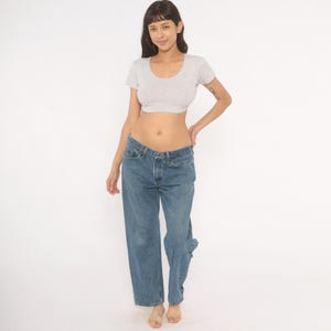 May include: A light gray, short-sleeved crop top with a scoop neck paired with blue denim jeans. The jeans have a relaxed fit and a classic five-pocket design. The model is standing in front of a white background.