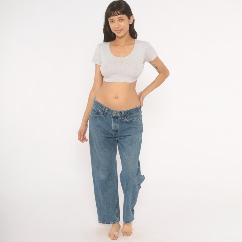 May include: A light gray, short-sleeved crop top with a scoop neck paired with blue denim jeans. The jeans have a relaxed fit and a classic five-pocket design. The model is standing in front of a white background.
