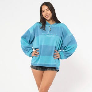 May include: A blue and turquoise striped long-sleeve shirt with a button-up collar. The shirt has a relaxed fit and features embroidered text above the buttons. The model is wearing black denim shorts.