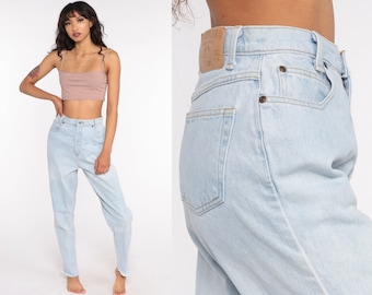 May include: Light blue high-waisted jeans with a faded wash and a raw hem. The jeans have a classic five-pocket design and a button and zipper closure. The model is wearing a light pink crop top.