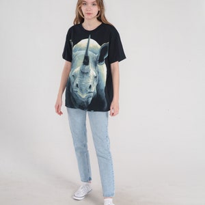 以下が含まれることがあります： 白いサイのグラフィックプリントが施された黒いTシャツ。サイは角を上に向けて視聴者の方を見ています。モデルは薄いブルーのジーンズと白いスニーカーを履いています。