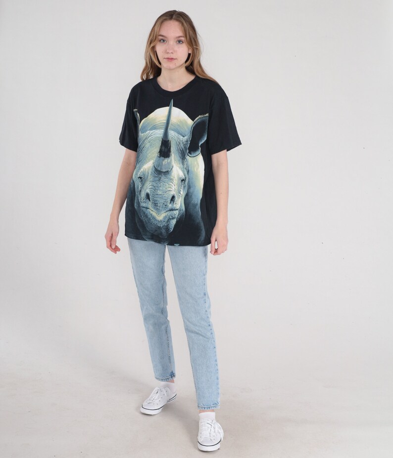 以下が含まれることがあります： 白いサイのグラフィックプリントが施された黒いTシャツ。サイは角を上に向けて視聴者の方を見ています。モデルは薄いブルーのジーンズと白いスニーカーを履いています。