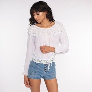 May include: A white long-sleeved blouse with a ruffled neckline and lace trim. The blouse has a cinched waist and is worn with denim shorts.