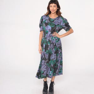May include: A floral print dress featuring a mix of green, purple, and black leaves. The dress has short sleeves and a midi length. The model is wearing black boots. The dress has a round neckline and a fitted waist.