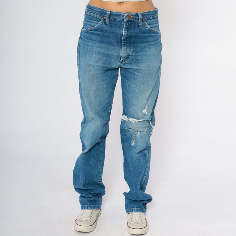 May include: Blue denim jeans with a distressed, ripped knee design. The jeans have a classic five-pocket style and a relaxed fit. The denim is faded in areas, giving them a vintage look. The jeans are worn with white canvas trainers.