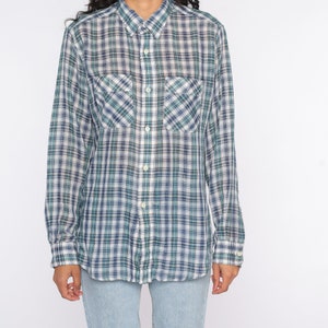 Op de afbeelding: Een shirt met lange mouwen met een blauw, groen en wit ruitpatroon. Het heeft twee borstzakken en een button-down kraag. De stof heeft een traditioneel ruitontwerp.