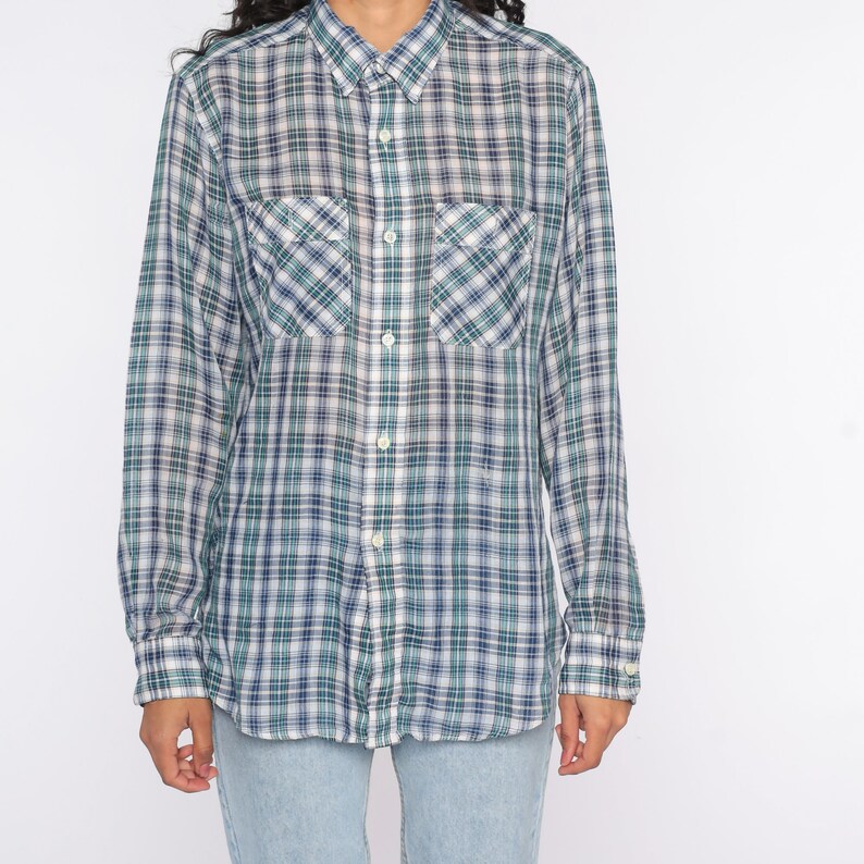 Op de afbeelding: Een shirt met lange mouwen met een blauw, groen en wit ruitpatroon. Het heeft twee borstzakken en een button-down kraag. De stof heeft een traditioneel ruitontwerp.