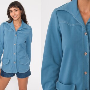 May include: A long-sleeved, light blue shirt with a pointed collar, button closure, and two patch pockets. The shirt has white stitching details.