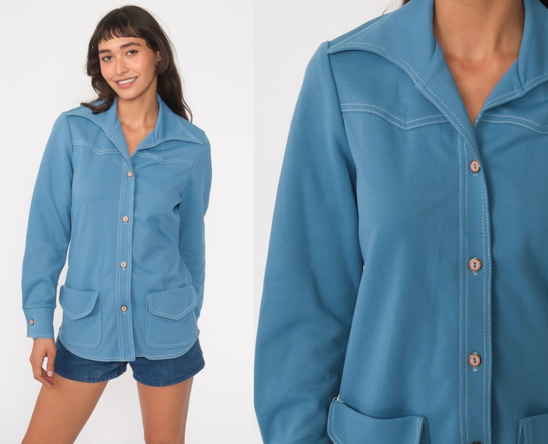 May include: A long-sleeved, light blue shirt with a pointed collar, button closure, and two patch pockets. The shirt has white stitching details.