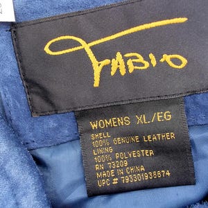 May include: Close-up of a blue suede garment with a black label. The label features the name "Fabio" in gold script and details including "WOMENS XL/EG", "100% GENUINE LEATHER", and "MADE IN CHINA".
