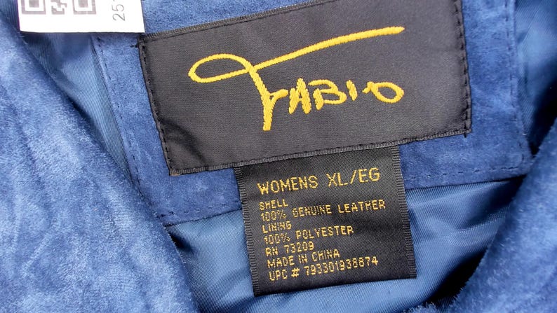 May include: Close-up of a blue suede garment with a black label. The label features the name "Fabio" in gold script and details including "WOMENS XL/EG", "100% GENUINE LEATHER", and "MADE IN CHINA".