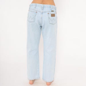 May include: Light blue denim jeans with a straight leg cut. The jeans feature the Wrangler logo on the back pockets and a brown leather patch. The jeans are a light wash and have a classic five-pocket design.