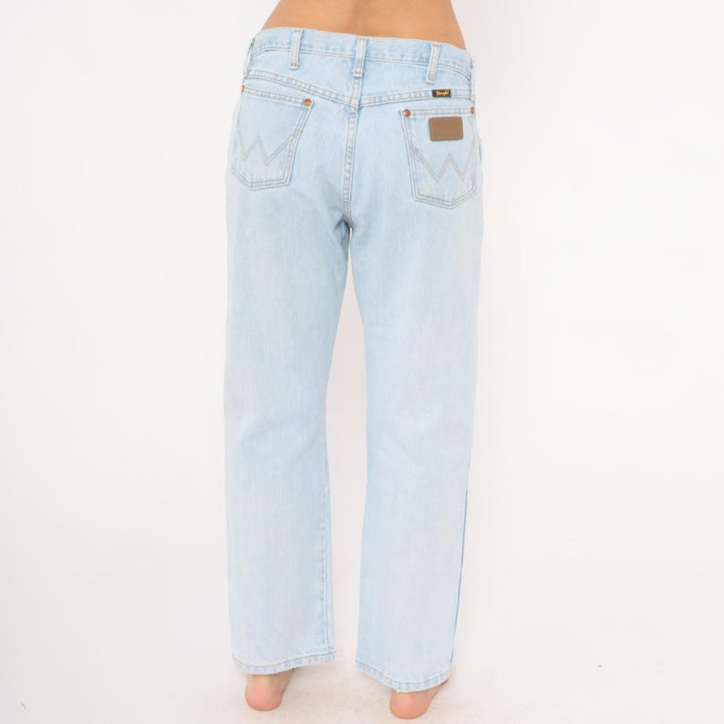 May include: Light blue denim jeans with a straight leg cut. The jeans feature the Wrangler logo on the back pockets and a brown leather patch. The jeans are a light wash and have a classic five-pocket design.