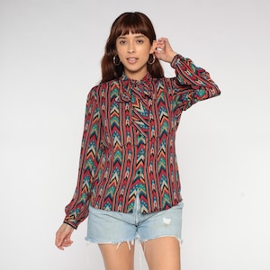 May include: A long-sleeved blouse with a colorful geometric pattern in red, teal, and yellow. The blouse features a bow tie neckline and is paired with denim shorts. The overall style is vintage and bohemian.
