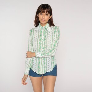 Puede incluir: Una camisa blanca de manga larga con un estampado floral verde y blanco. La camisa tiene cuello y cierre de botones en la parte delantera.