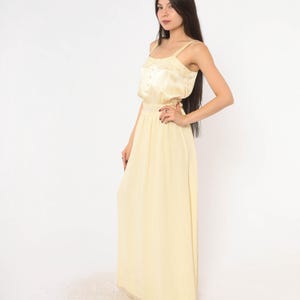 May include: A floor-length pale yellow sleeveless dress featuring a fitted bodice with a row of small buttons down the centre, wide shoulder straps, and a long, flowing skirt that reaches the floor, crafted from a smooth, lightweight fabric.