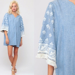 May include: A light blue mini dress featuring a V-neckline and wide, elbow-length sleeves. The sleeves are decorated with white floral embroidery, a wide lace band, and a fringe trim along the edges. The fabric has a visible, textured weave.