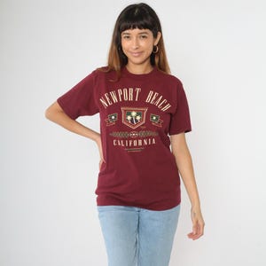 May include: A maroon t-shirt with the text "NEWPORT BEACH CALIFORNIA" in a crest design. The crest features palm trees and banners. The shirt is paired with light blue jeans.