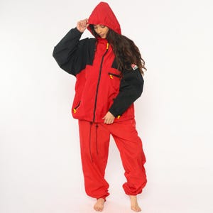 May include: Red and black hooded jacket and matching pants. The jacket has a red body with black sleeves and a hood. Yellow accents are on the zippers and pockets. The pants are red with a drawstring waist. A vintage outdoor sports outfit.