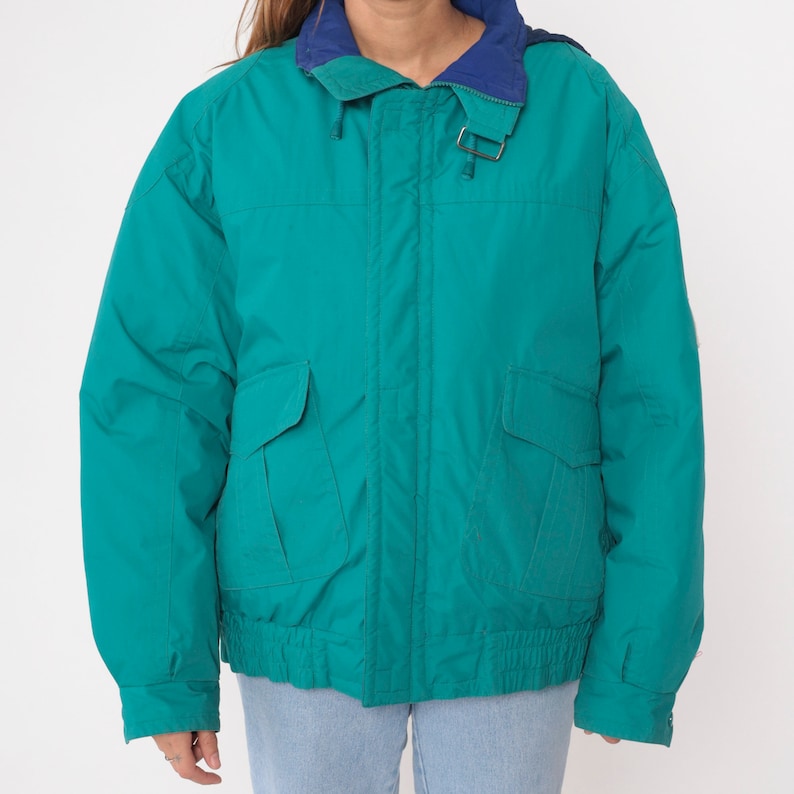 K&ouml;nnte beinhalten: Eine tealblaue Jacke mit Rei&szlig;verschluss und zwei Fronttaschen. Die Jacke hat ein blaues Futter am Kragen und eine Kapuze mit Kordelzug.