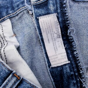 Puede incluir: Primer plano de unos vaqueros azules con una etiqueta blanca. La etiqueta dice "Wrangler" e incluye informaci&oacute;n sobre la talla. Los vaqueros tienen costuras visibles y un bot&oacute;n met&aacute;lico.