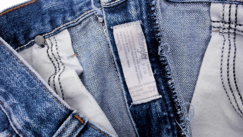 Puede incluir: Primer plano de unos vaqueros azules con una etiqueta blanca. La etiqueta dice "Wrangler" e incluye informaci&oacute;n sobre la talla. Los vaqueros tienen costuras visibles y un bot&oacute;n met&aacute;lico.