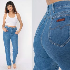 Op de afbeelding: Een paar blauwe denim jeans met een hoge taille en een klassieke rechte pijp. De jeans heeft een bruin leren patch met het woord "Wrangler" op de rechter achterzak.
