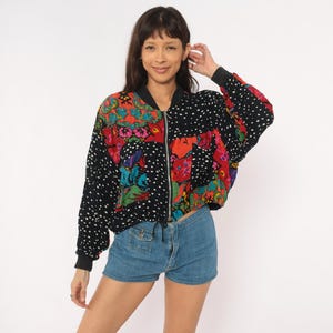 May include: A long-sleeve bomber jacket featuring a mix of black fabric with white polka dots and panels of vibrant floral patterns in red, green, and blue. The jacket has a central silver zipper, a black ribbed collar, and black ribbed cuffs.