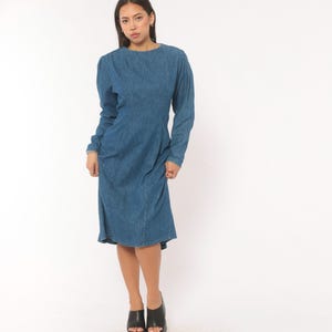 Puede incluir: Un vestido de mezclilla azul de manga larga con cuello redondo, hombros estructurados y un corpi&ntilde;o ajustado que se transforma en una falda ligeramente acampanada. La prenda presenta costuras verticales visibles en la parte delantera.