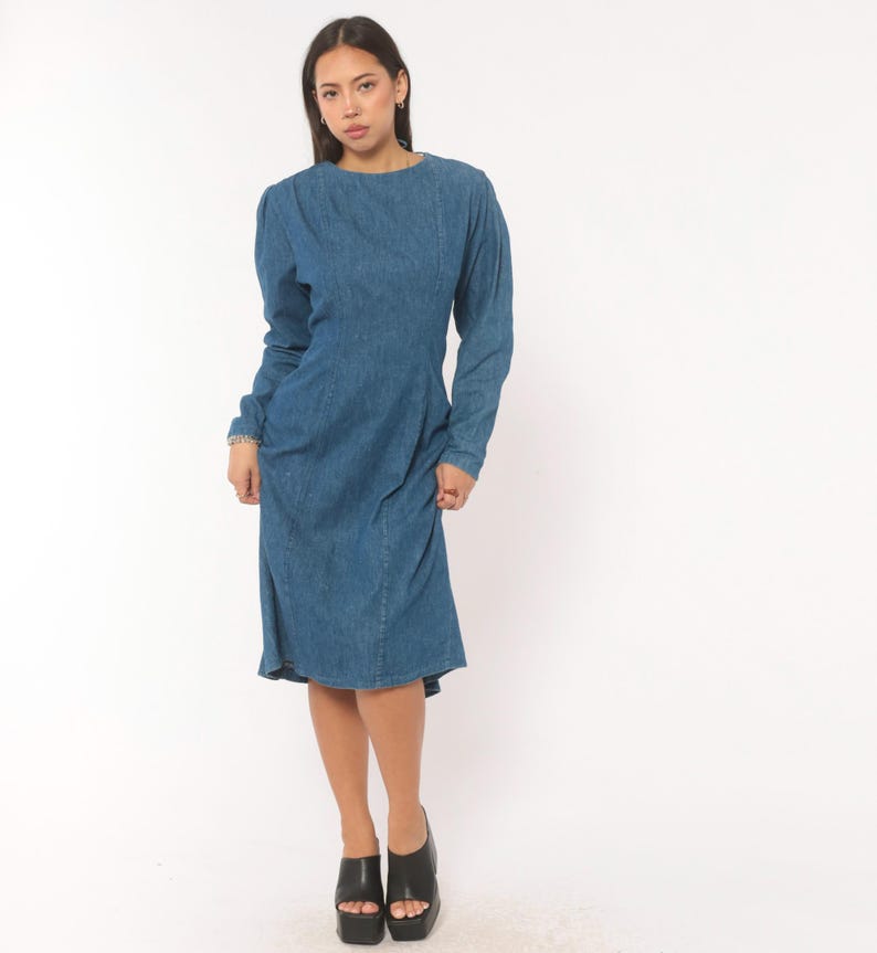 Puede incluir: Un vestido de mezclilla azul de manga larga con cuello redondo, hombros estructurados y un corpi&ntilde;o ajustado que se transforma en una falda ligeramente acampanada. La prenda presenta costuras verticales visibles en la parte delantera.