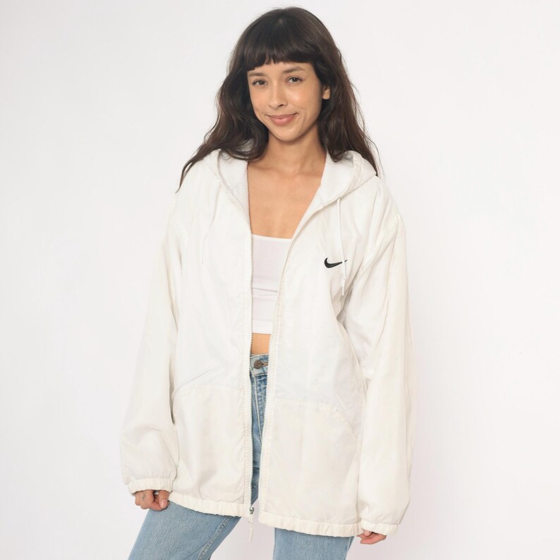 Puede incluir: Chaqueta blanca con capucha y logotipo negro de Nike en el pecho. La chaqueta tiene cremallera completa y capucha con cord&oacute;n. La modelo lleva vaqueros azules y una camiseta blanca.