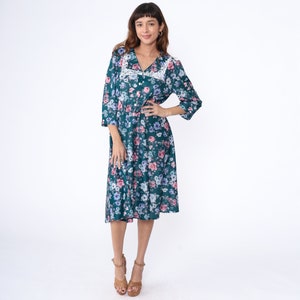 K&ouml;nnte beinhalten: Ein blaues Midi-Kleid in Teal mit einem wei&szlig;en Spitzenkragen und einem Blumendruck mit rosa, blauen und wei&szlig;en Blumen. Das Kleid hat einen Knopfleisten vorne und 3/4 &Auml;rmel.