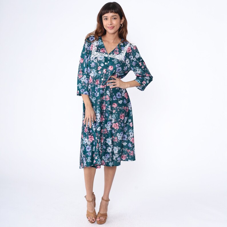 K&ouml;nnte beinhalten: Ein blaues Midi-Kleid in Teal mit einem wei&szlig;en Spitzenkragen und einem Blumendruck mit rosa, blauen und wei&szlig;en Blumen. Das Kleid hat einen Knopfleisten vorne und 3/4 &Auml;rmel.