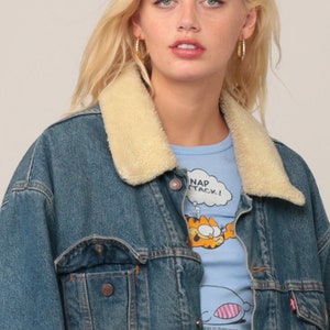 Puede incluir: Una chaqueta de mezclilla azul con cuello de piel sint&eacute;tica color crema, con botones de metal y un bolsillo en el pecho. Debajo, una camiseta azul claro muestra un gr&aacute;fico del personaje de dibujos animados Garfield con el texto NAP ATTACK.