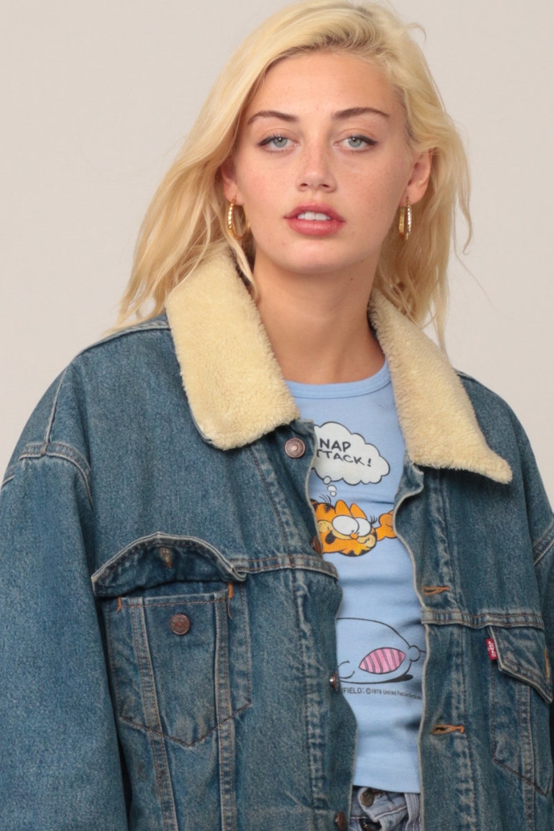 Puede incluir: Una chaqueta de mezclilla azul con cuello de piel sint&eacute;tica color crema, con botones de metal y un bolsillo en el pecho. Debajo, una camiseta azul claro muestra un gr&aacute;fico del personaje de dibujos animados Garfield con el texto NAP ATTACK.