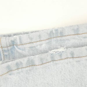 May include: Close-up of light blue denim jeans. The image shows the waistband and a portion of the leg, with visible stitching and a small tear. The fabric has a faded appearance.