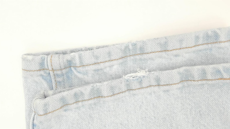 May include: Close-up of light blue denim jeans. The image shows the waistband and a portion of the leg, with visible stitching and a small tear. The fabric has a faded appearance.
