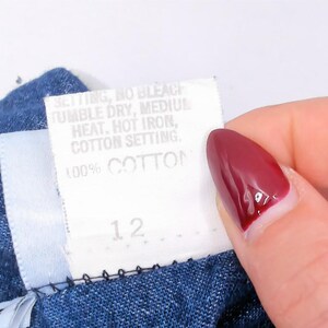 Puede incluir: Una prenda de tela azul con una etiqueta de cuidado blanca que muestra el texto: SETTING, NO BLEACH, TUMBLE DRY, MEDIUM HEAT, HOT IRON, COTTON SETTING, 100% COTTON y el n&uacute;mero 12. La etiqueta proporciona instrucciones de lavado para el art&iacute;culo.