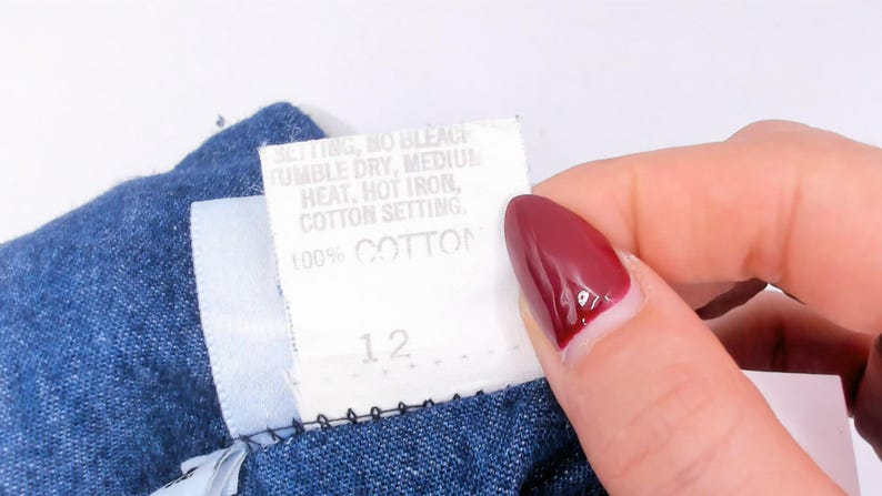 Puede incluir: Una prenda de tela azul con una etiqueta de cuidado blanca que muestra el texto: SETTING, NO BLEACH, TUMBLE DRY, MEDIUM HEAT, HOT IRON, COTTON SETTING, 100% COTTON y el n&uacute;mero 12. La etiqueta proporciona instrucciones de lavado para el art&iacute;culo.