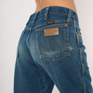 May include: Close-up of blue denim jeans with the Wrangler logo on the back pocket. The jeans have a classic fit with copper rivets and a brown leather patch. The denim has a faded, worn look.