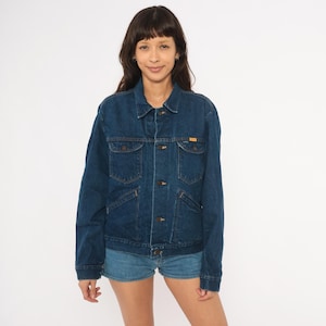 May include: A dark blue denim jacket featuring a classic collar, a front button closure, two chest pockets with buttoned flaps, and two lower side pockets. The jacket has long sleeves with buttoned cuffs and a straight hemline, constructed from durable cotton denim fabric.