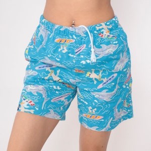 K&ouml;nnte beinhalten: Blaue Badeshorts mit einem Muster aus Haien, Surfern und Wellen. Die Shorts haben einen Kordelzug.