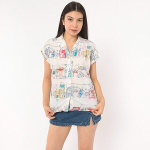 May include: A short-sleeved button-down shirt in white featuring a colorful hand-drawn style print of various buildings and architectural structures in shades of blue, red, yellow, and green, paired with a dark blue denim mini skirt.