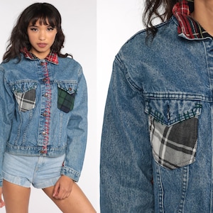 May include: A light blue denim jacket with a red stitched seam down the center and plaid accents. The jacket features a plaid collar and two front pockets with plaid patches. The jacket has brown buttons and long sleeves.