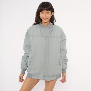 May include: A light gray bomber jacket featuring a ribbed collar, ribbed cuffs, and a ribbed hem. The jacket has a front zipper closure concealed by a placket, long sleeves, and two side pockets. The fabric has a smooth, matte finish throughout.