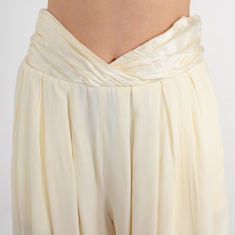 May include: Cream-colored high-waisted pants with a draped waistband. The pants have a pleated front and a wide-leg design, made of a soft, flowing fabric. The neutral color makes them versatile for various outfits.