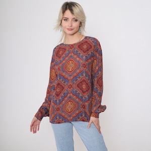 May include: A long-sleeved, loose-fitting blouse with a colorful paisley print in shades of orange, red, blue, and purple. The blouse has a round neckline and a relaxed fit.